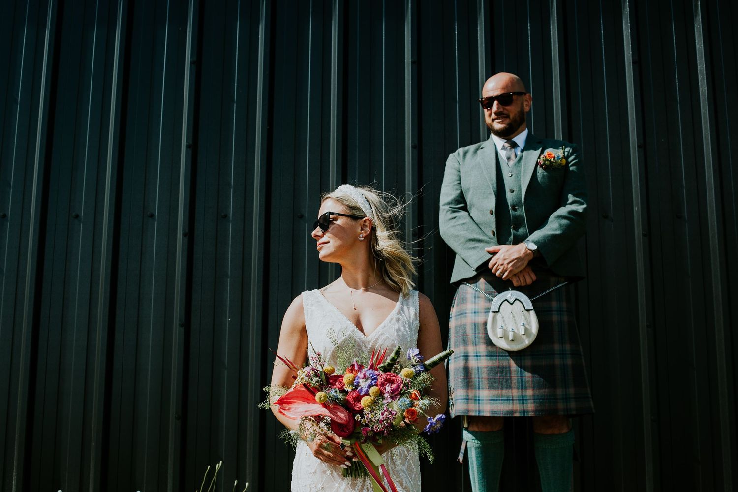 Stylish wedding couple in sunglasses pose against dark metal wall wearing Scottish formal wear.