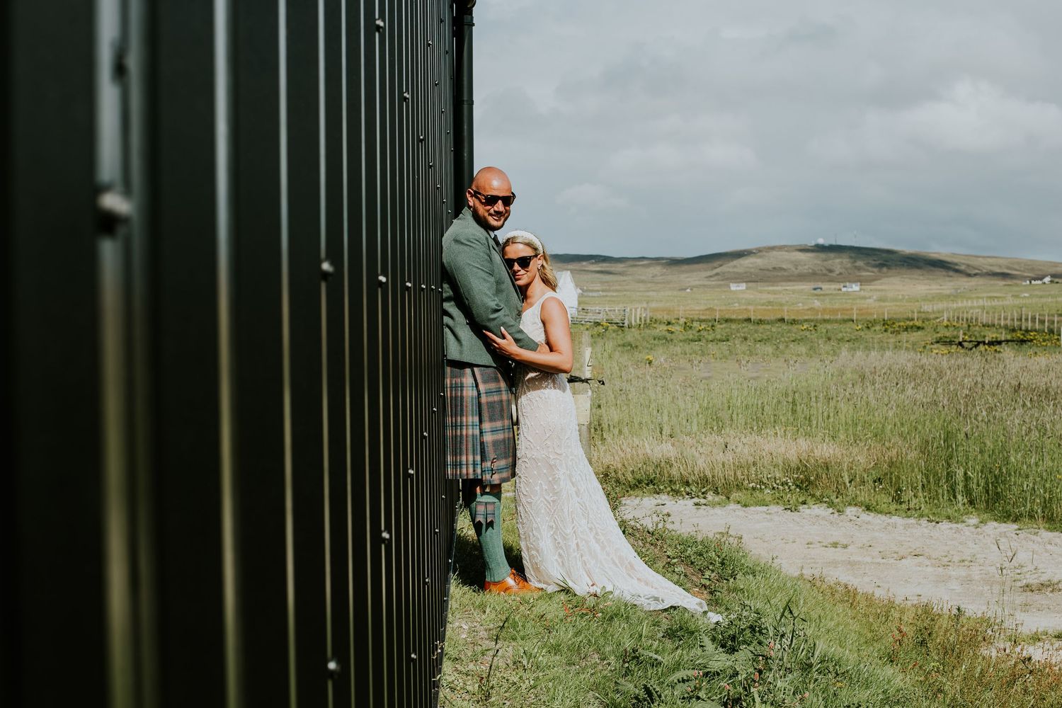A wedding couple poses playfully against a dark wall with rolling hills in the background.