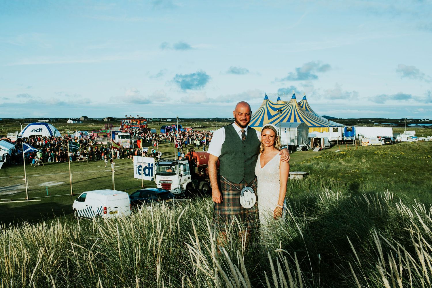 A couple stands in a coastal landscape with beach grass and tents during an outdoor event under blue skies.
