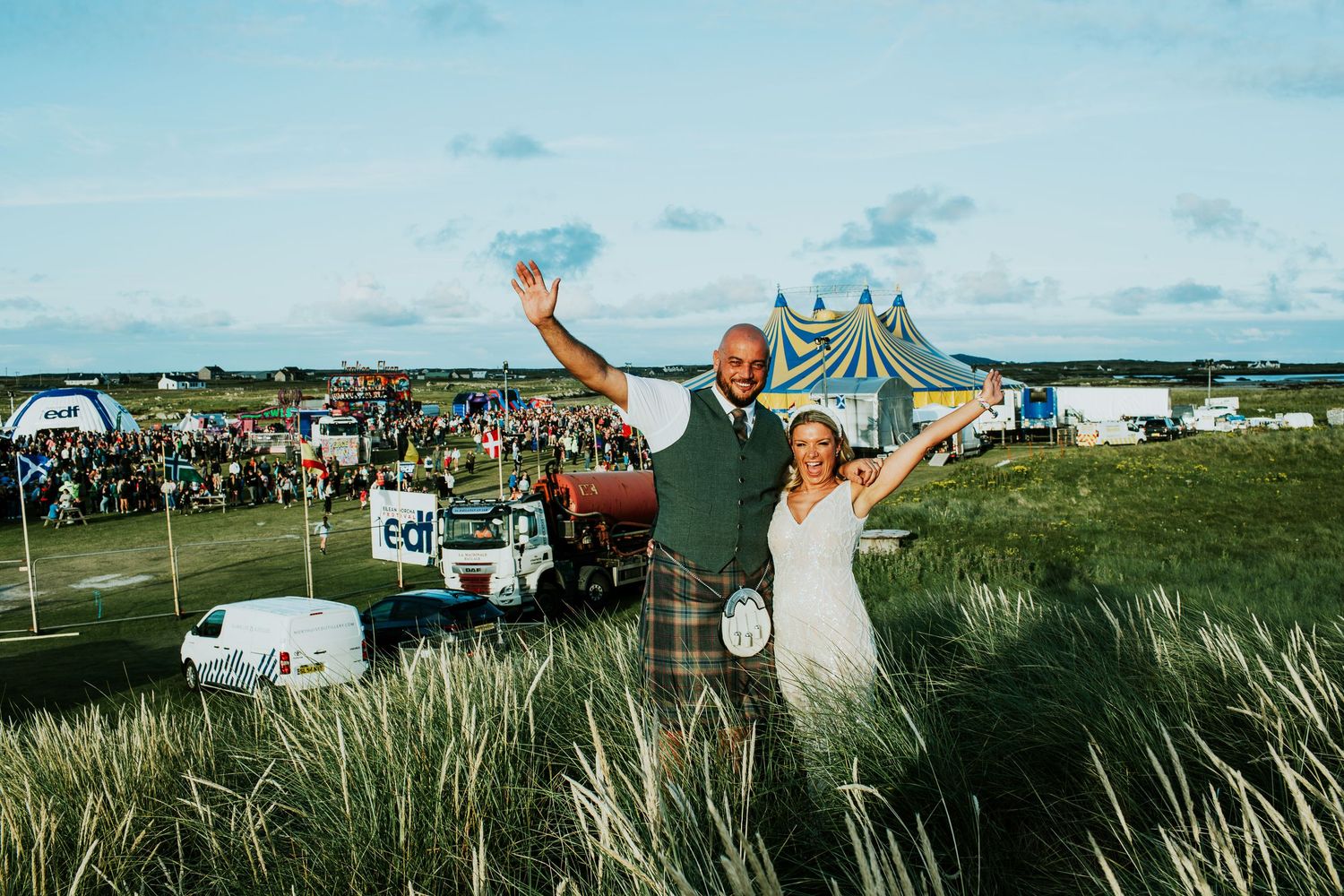 A couple celebrates outdoors at a festival with a yellow and blue striped tent and parked vehicles in the background.