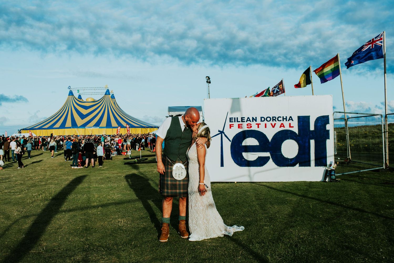 A couple poses in front of a yellow and blue striped tent with various flags and an EDI banner at an outdoor event.