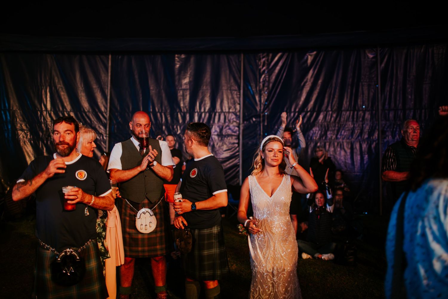 A group celebrates at a wedding reception with men in kilts and festive dancing under dramatic lighting.