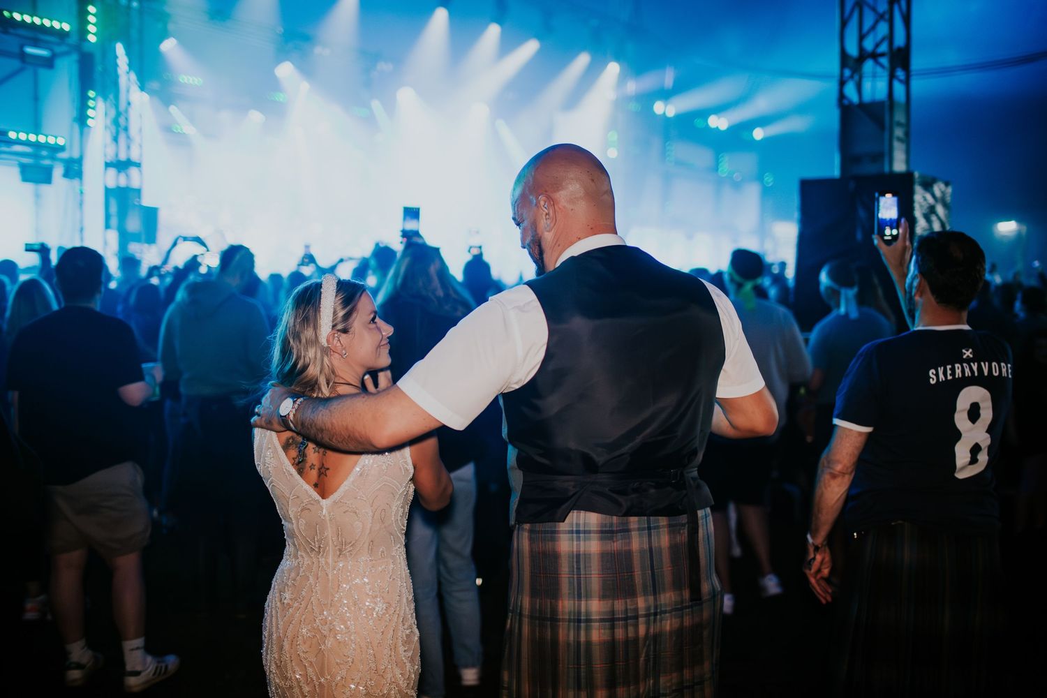 A couple in formal attire, with a person wearing a kilt, dancing at an event with blue stage lighting.