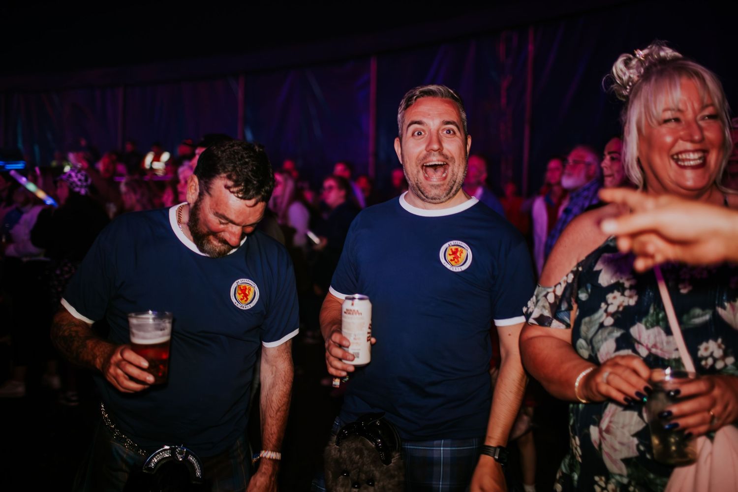 Scottish football fans in blue jerseys holding drinks at a crowded indoor venue during a match gathering.