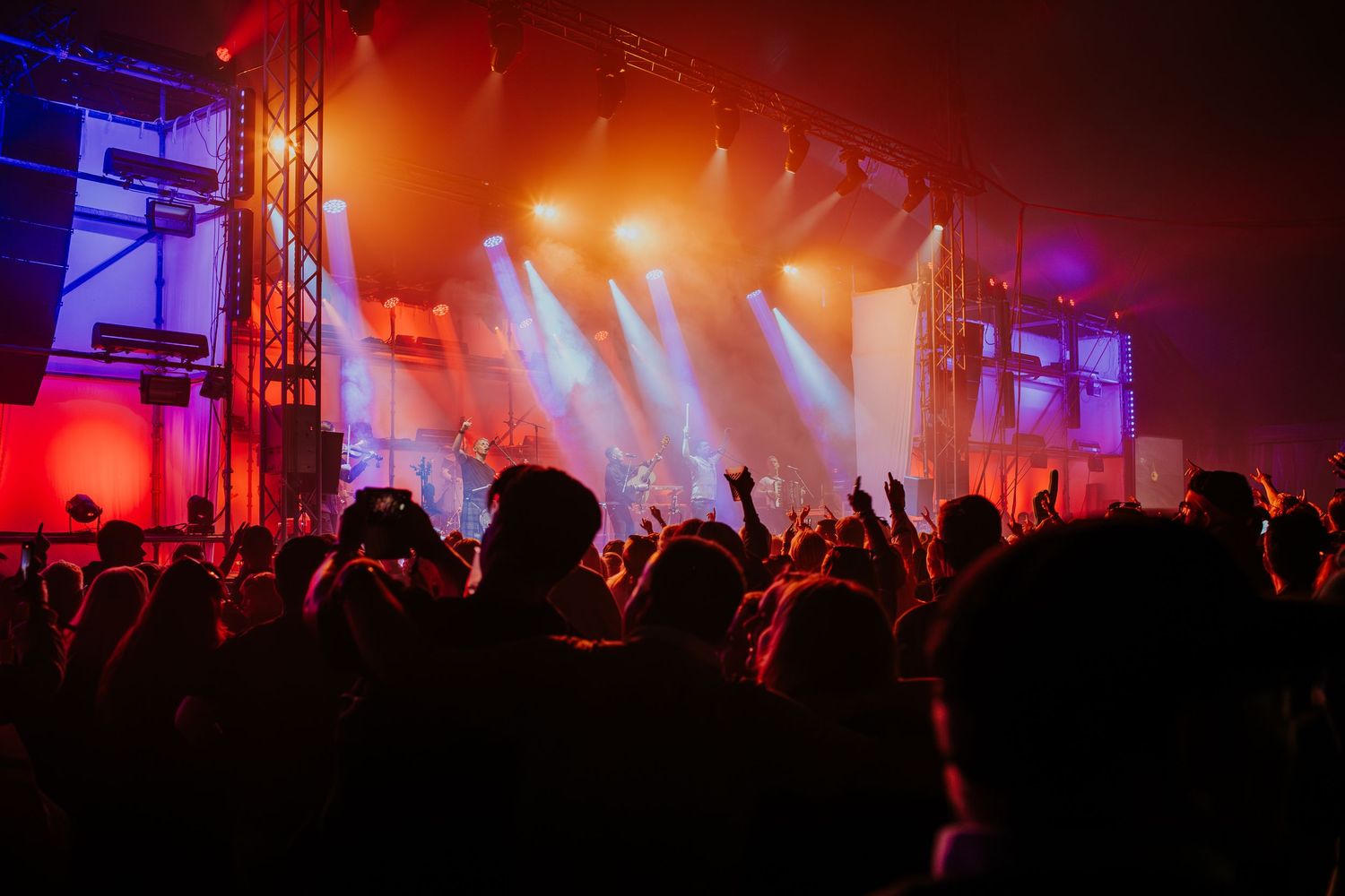 Concert crowd silhouettes with raised hands against red stage lighting and fog at a live music performance.