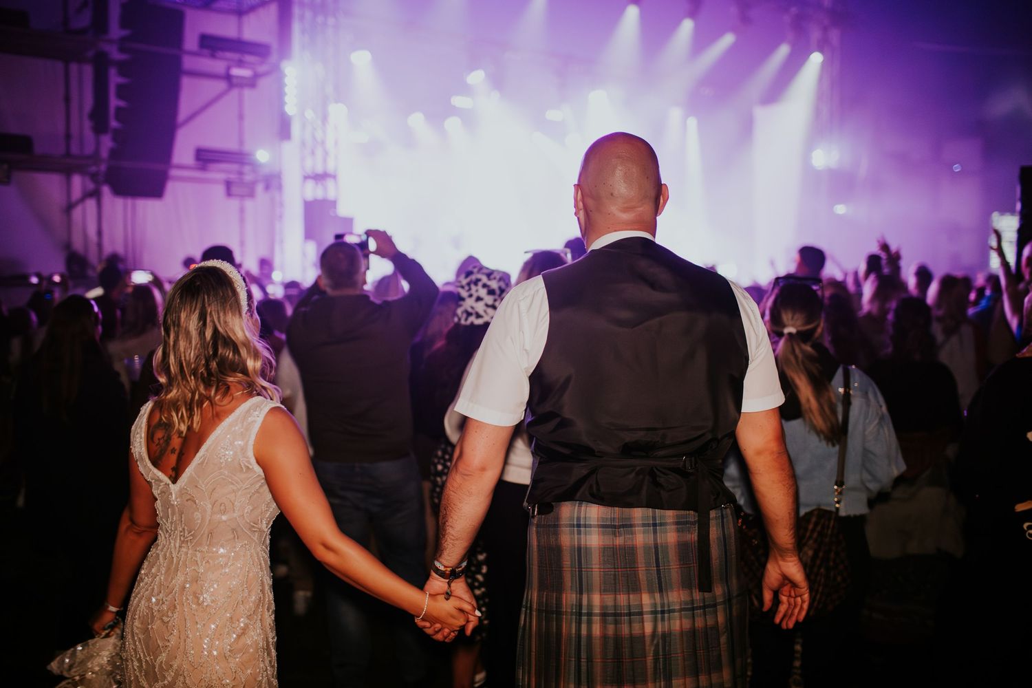A couple holds hands while watching a purple-lit concert stage at an evening music festival.