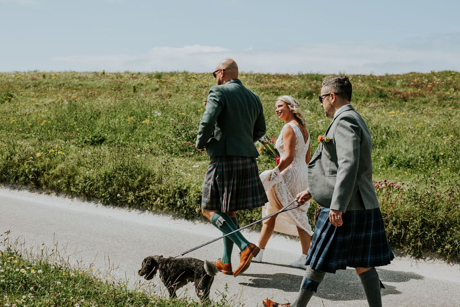 Three people in kilts walking a small dog on a paved path through green countryside on a sunny day.