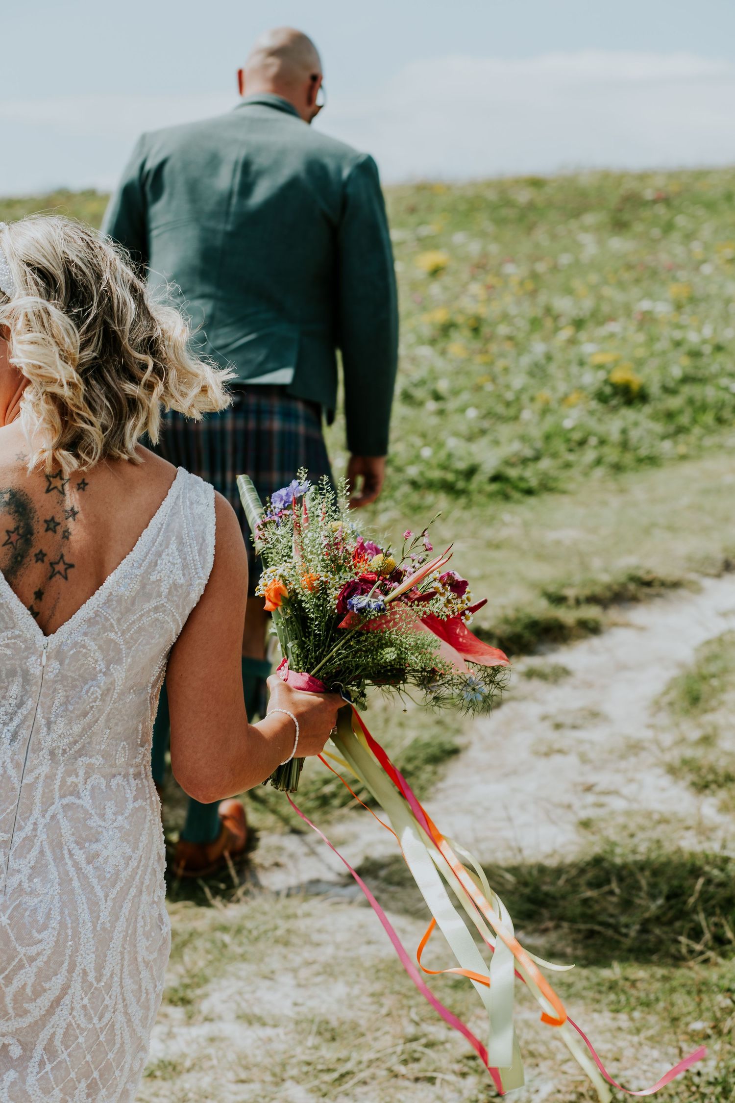 Wedding couple walking on coastal path with bride carrying colorful wildflower bouquet with ribbons.