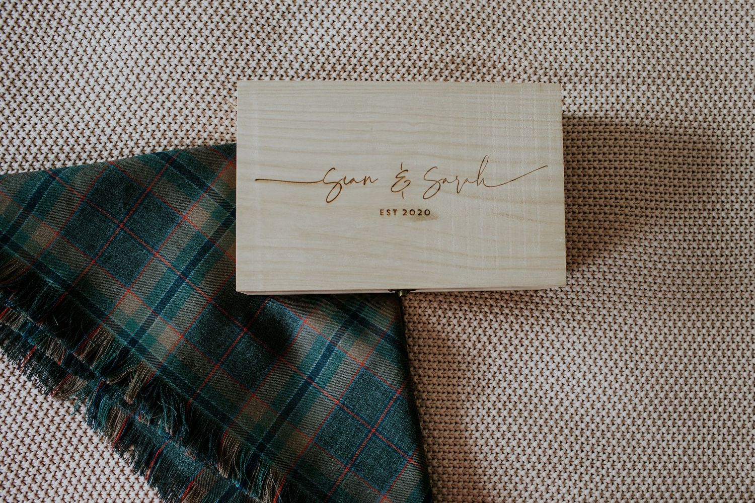 Small wooden box with cursive engraving displayed next to plaid fabric on textured gray surface.