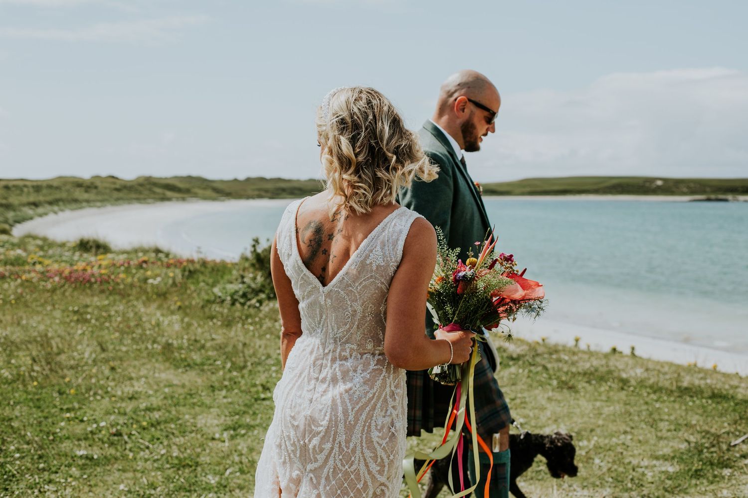 A couple stands on a grassy oceanfront cliff during their wedding ceremony, with the bride holding a colorful bouquet.