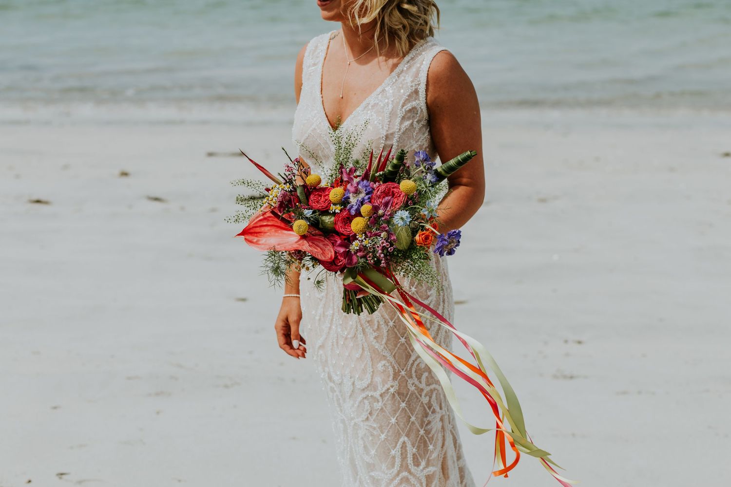 Person in lace wedding dress holds vibrant tropical bouquet with red flowers and colorful ribbons on sandy beach.