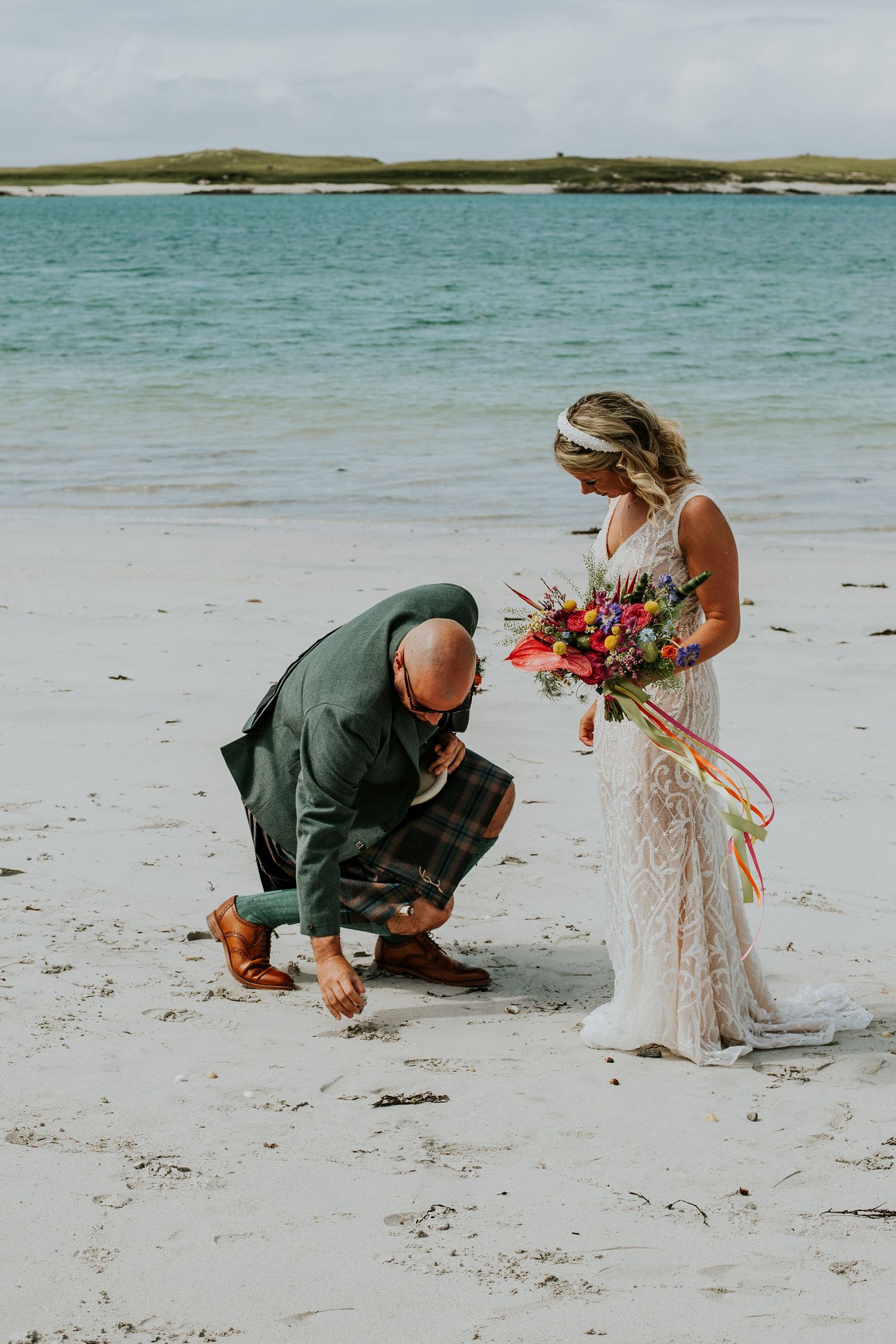 A couple on a tropical beach during a wedding ceremony as one adjusts the others dress against turquoise waters.
