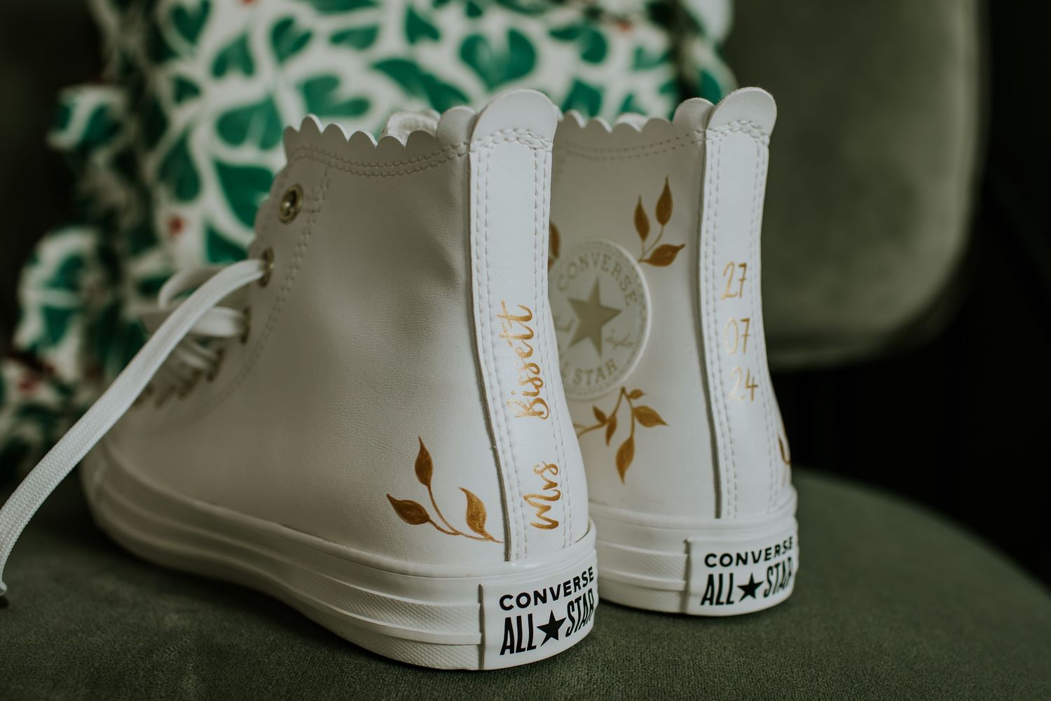 White leather high-top Converse sneaker with delicate gold floral design details sits against green background.