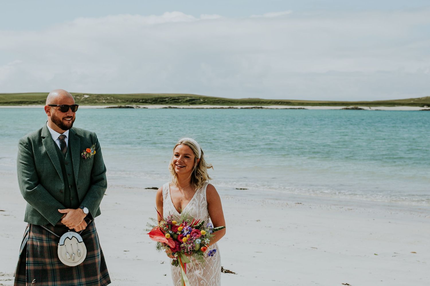 Two people on a scenic beach with turquoise water, with one wearing a traditional Scottish kilt and sporran.