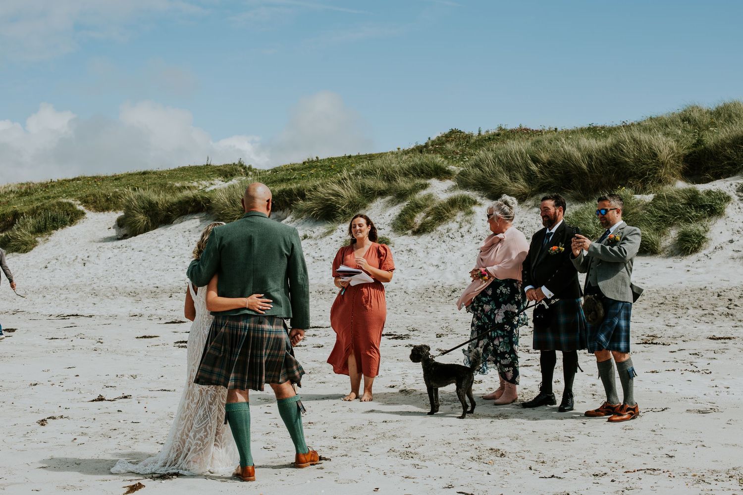 Five people and a dog gather on a sandy beach near grassy dunes on a cloudy day.