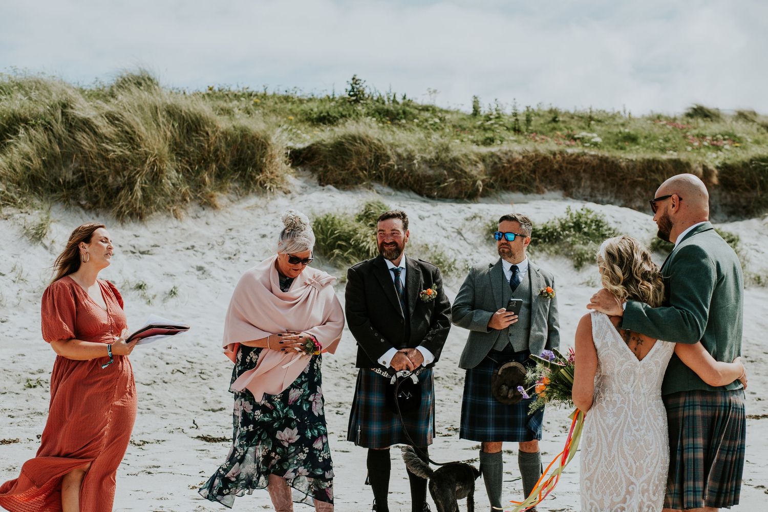 A beach wedding ceremony with guests wearing traditional Scottish kilts on a sandy shore.