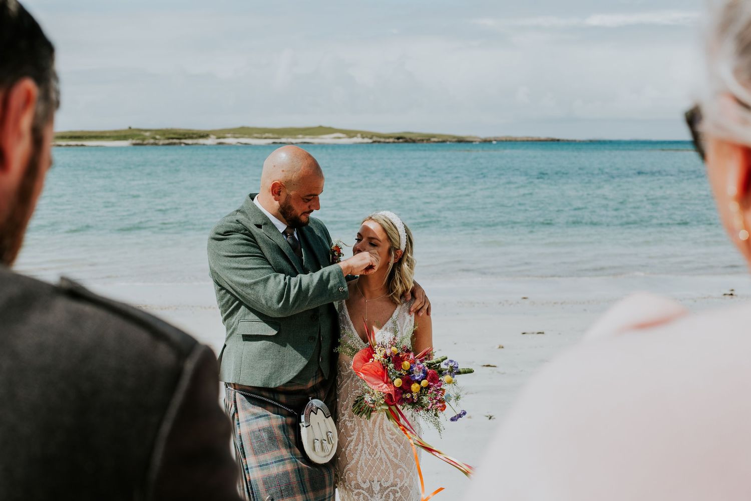 A romantic beach wedding ceremony takes place on a scenic shoreline with turquoise waters and green hills in the distance.