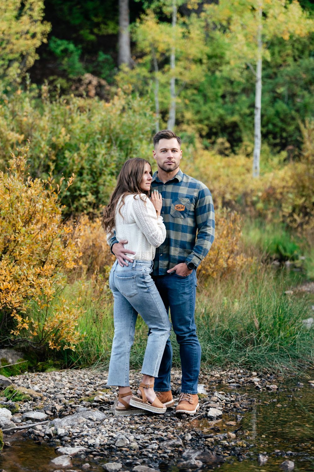 A couple stands together on rocky ground near a stream surrounded by autumn foliage.