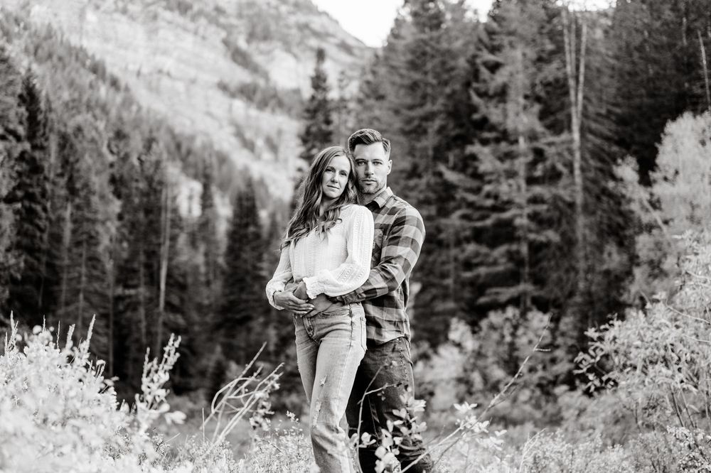 Black and white photo of couple embracing in mountain valley with evergreen forest backdrop.
