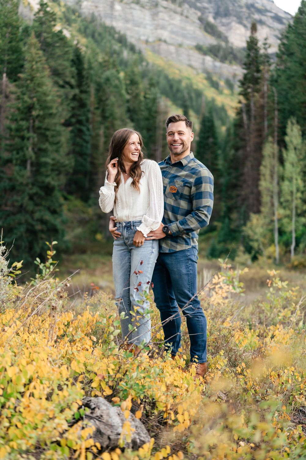 A couple stands together in a mountain meadow filled with autumn foliage and evergreen trees.