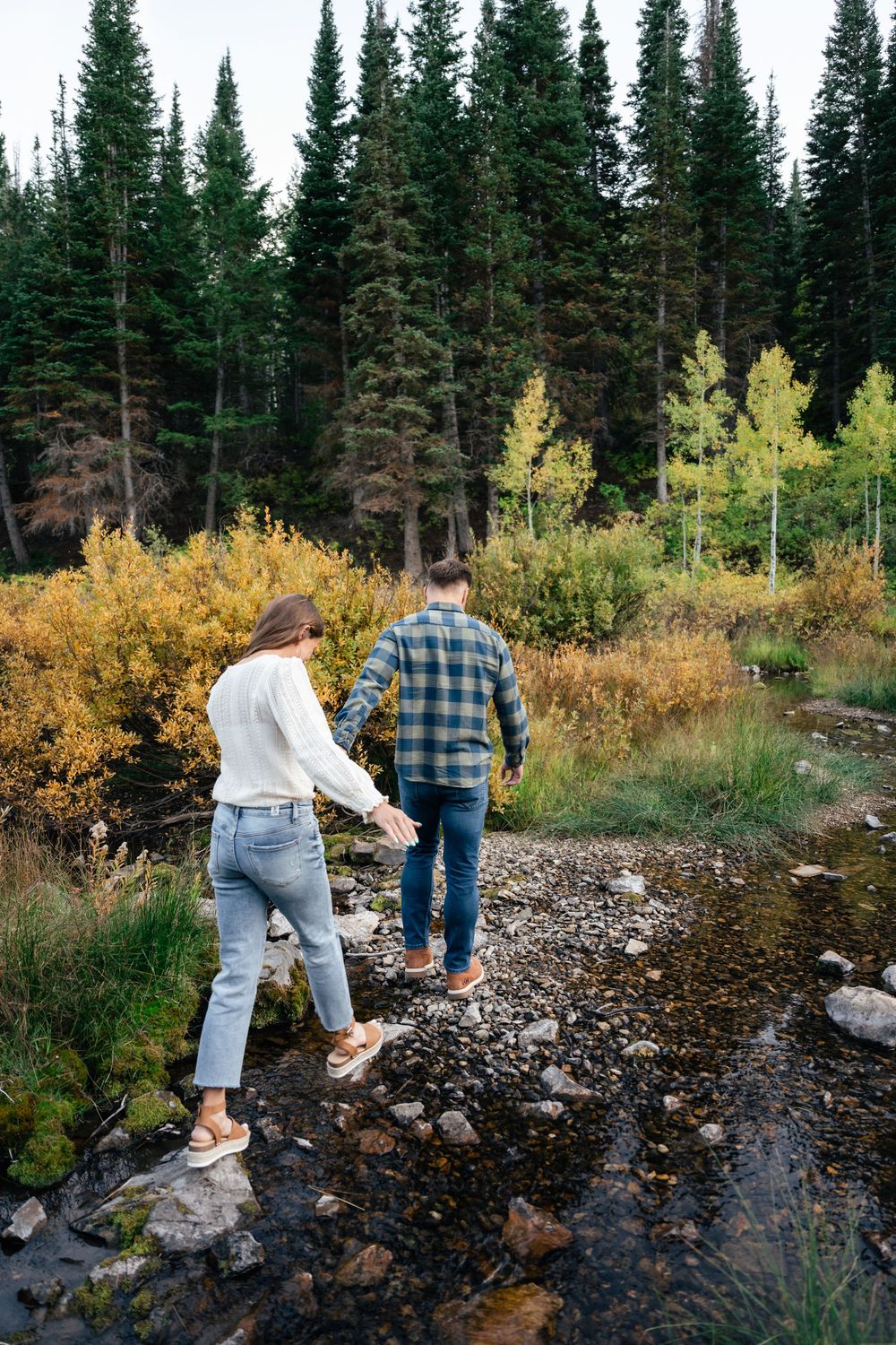 Couple crossing rocky stream together surrounded by evergreen forest and autumn foliage in mountains.