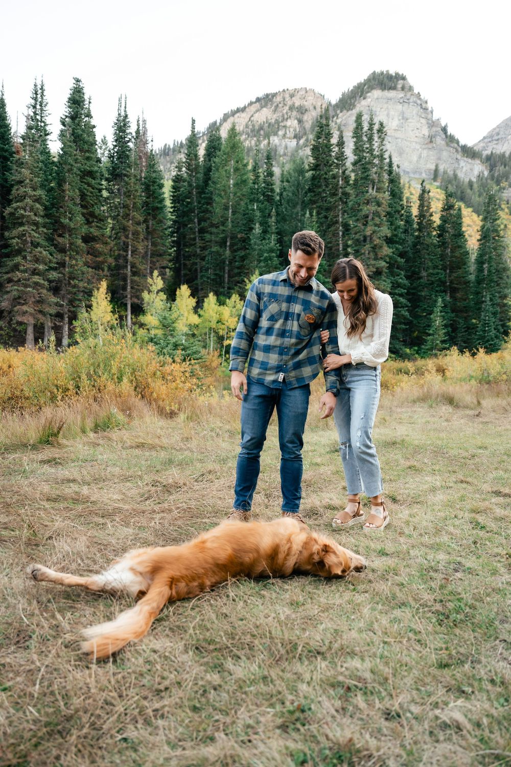 A couple laughs together in the mountains while their golden retriever rests on the ground nearby.