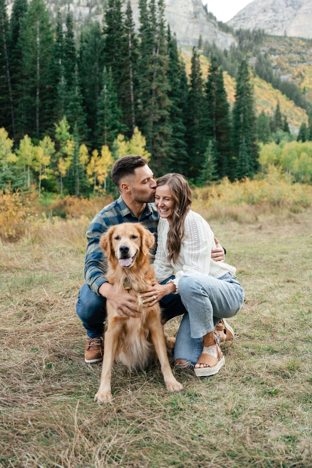 A happy couple poses with their Golden Retriever dog against a backdrop of mountain scenery and fall colors.