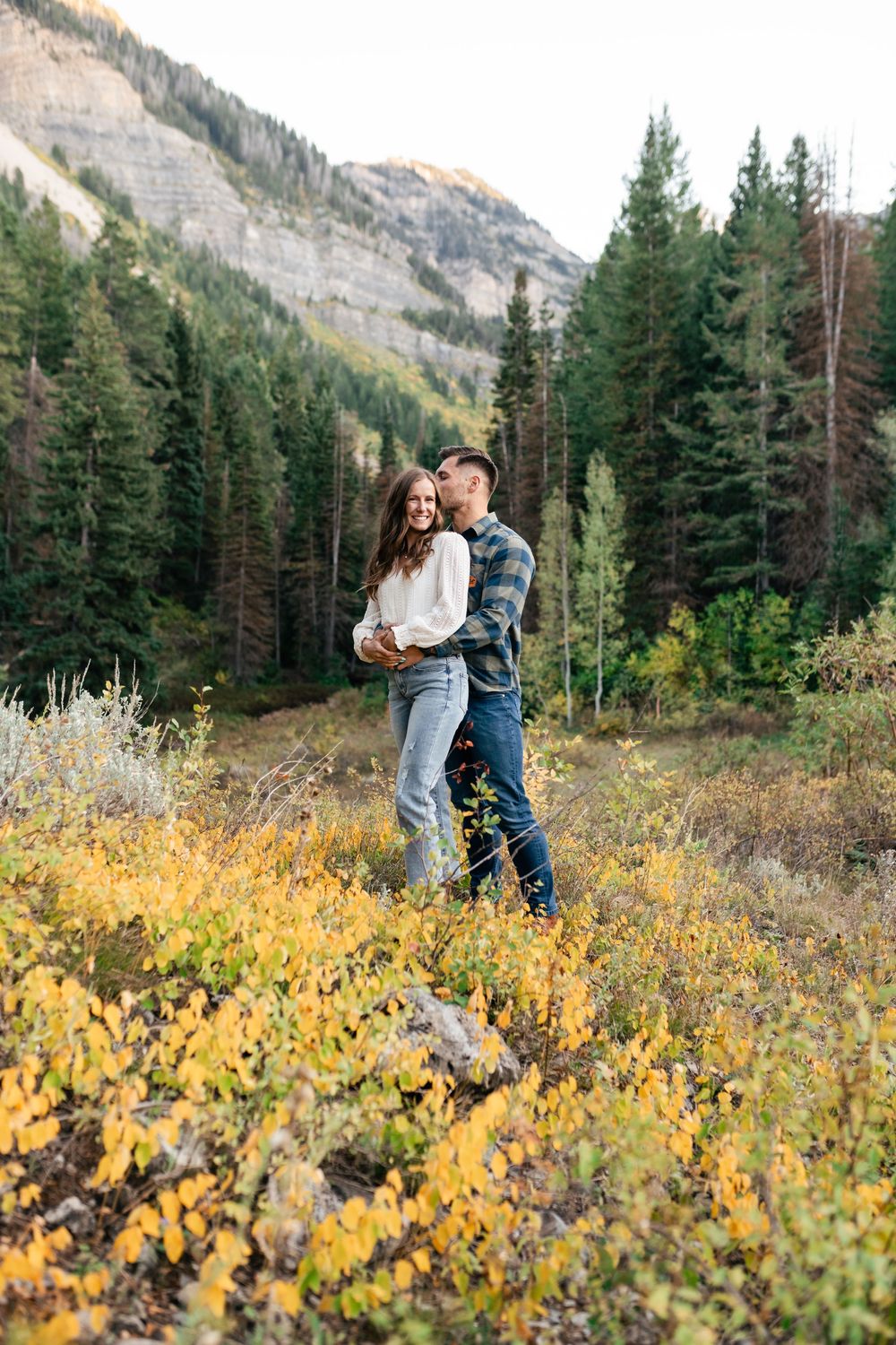 A romantic couple embraces while standing in mountain wilderness surrounded by yellow wildflowers and evergreen trees.