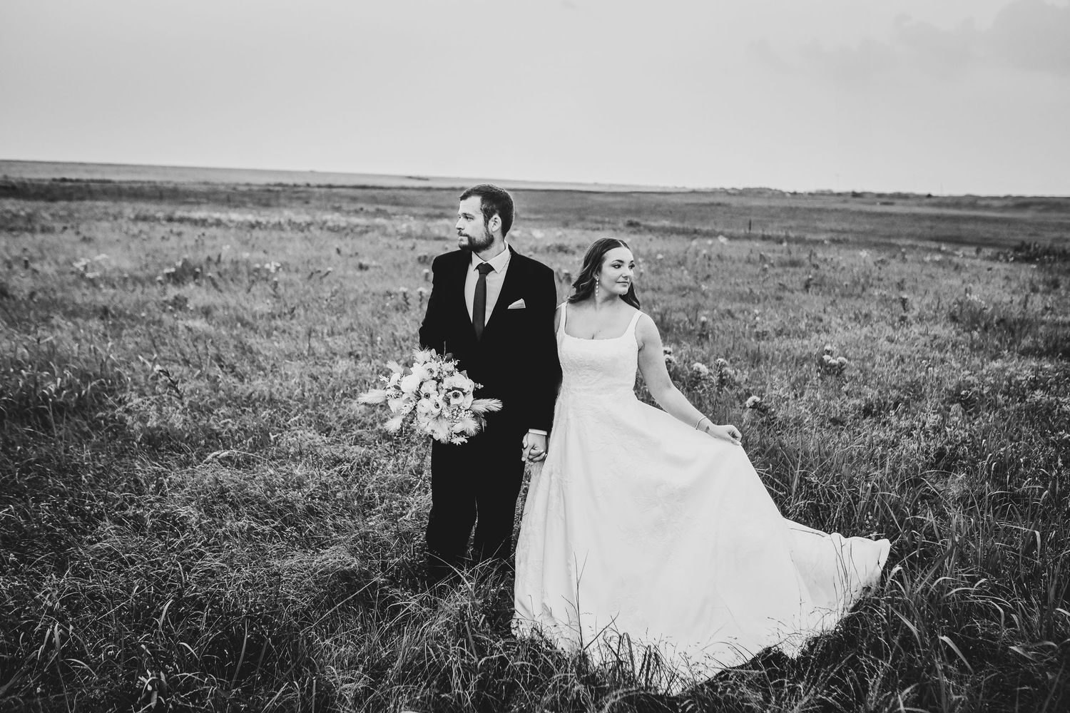 A black and white romantic portrait of a bride and groom holding hands in a field of golden grass outside of their dallas wedding venue
