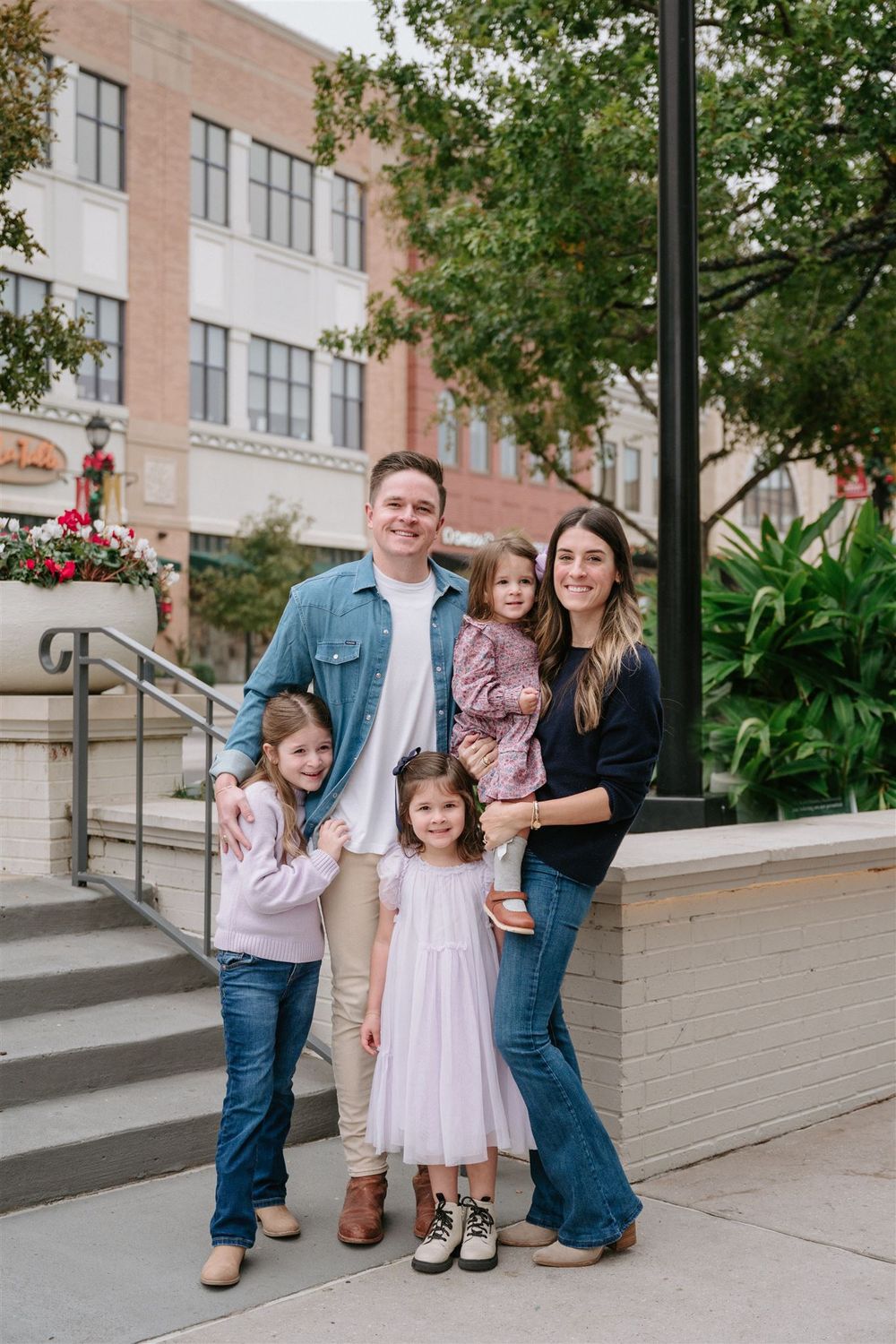 A family of five poses together on urban steps with brick buildings and greenery in the background.