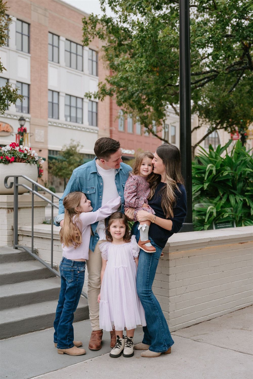 A family stands together on steps outside a brick building with trees and greenery in the background.
