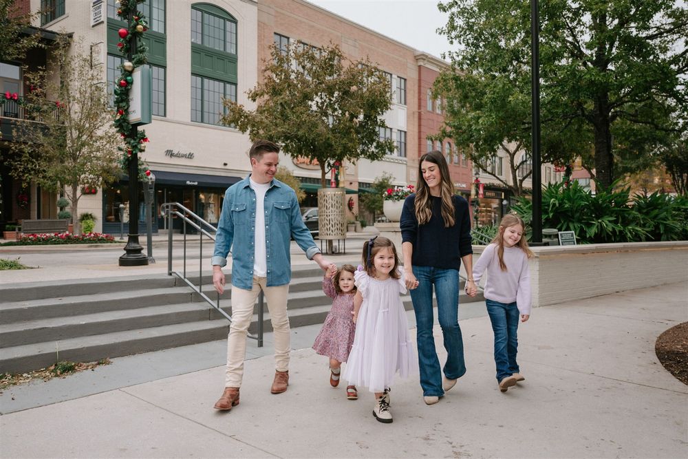 A family of five strolls together on a city sidewalk past modern storefronts and trees on a sunny day.