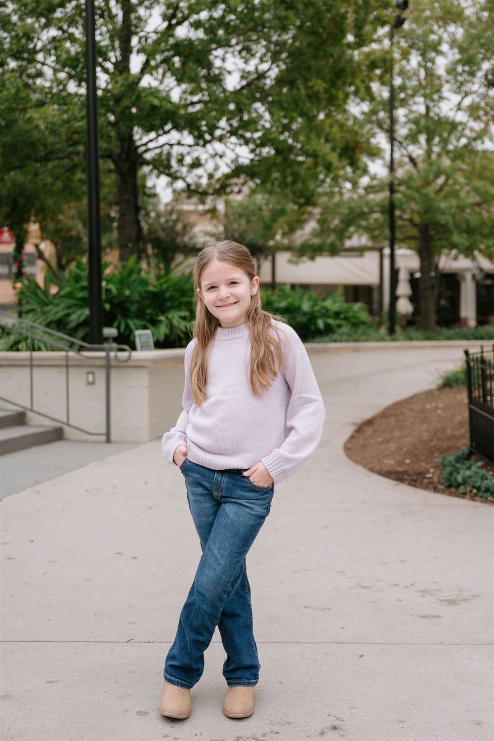 A young person wearing a light pink sweater and blue jeans poses casually on a curved concrete pathway in an outdoor setting.