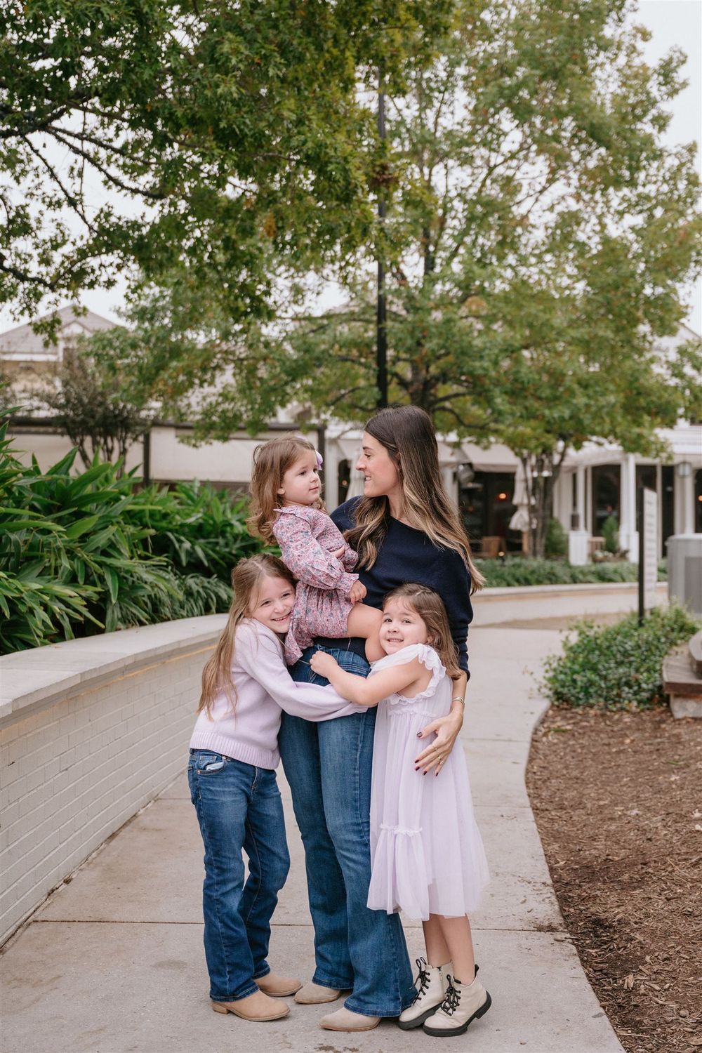 Woman wearing denim and black sweater stands on outdoor pathway with three children in casual attire.