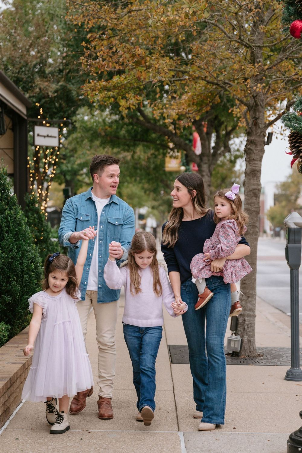 A family walks together down a tree-lined sidewalk on an autumn day wearing casual clothing.
