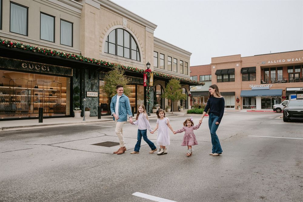A family group holds hands and dances together on a quiet street with retail storefronts in the background.