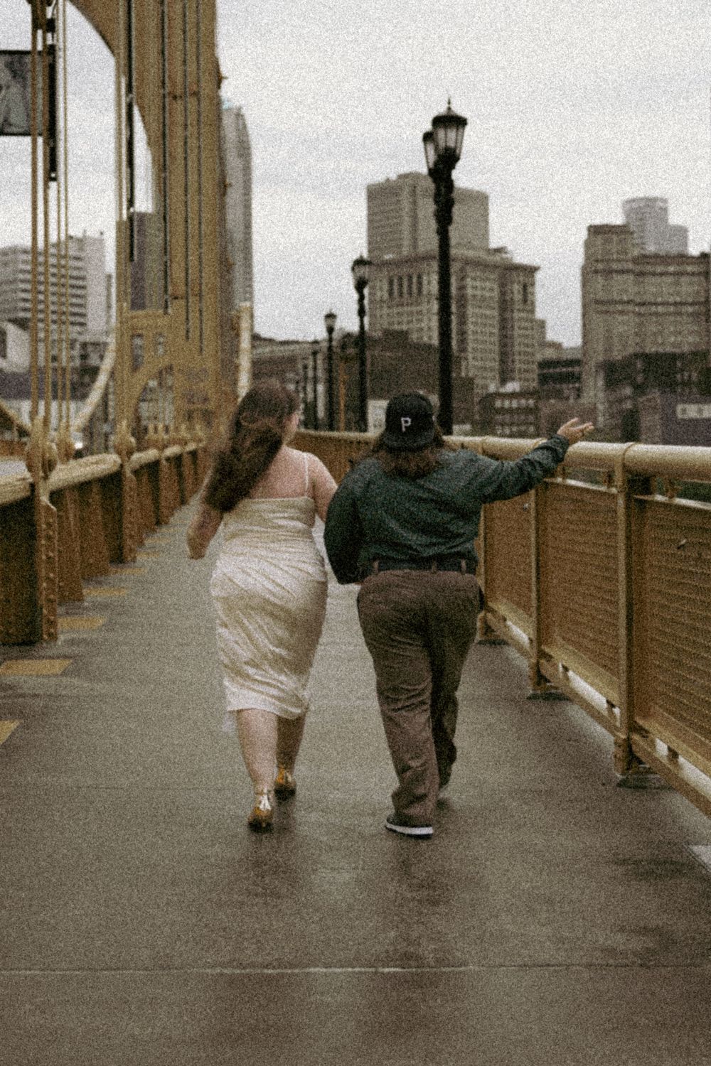 Two people walking hand in hand on a city bridge in casual attire on an overcast day with urban buildings in background.