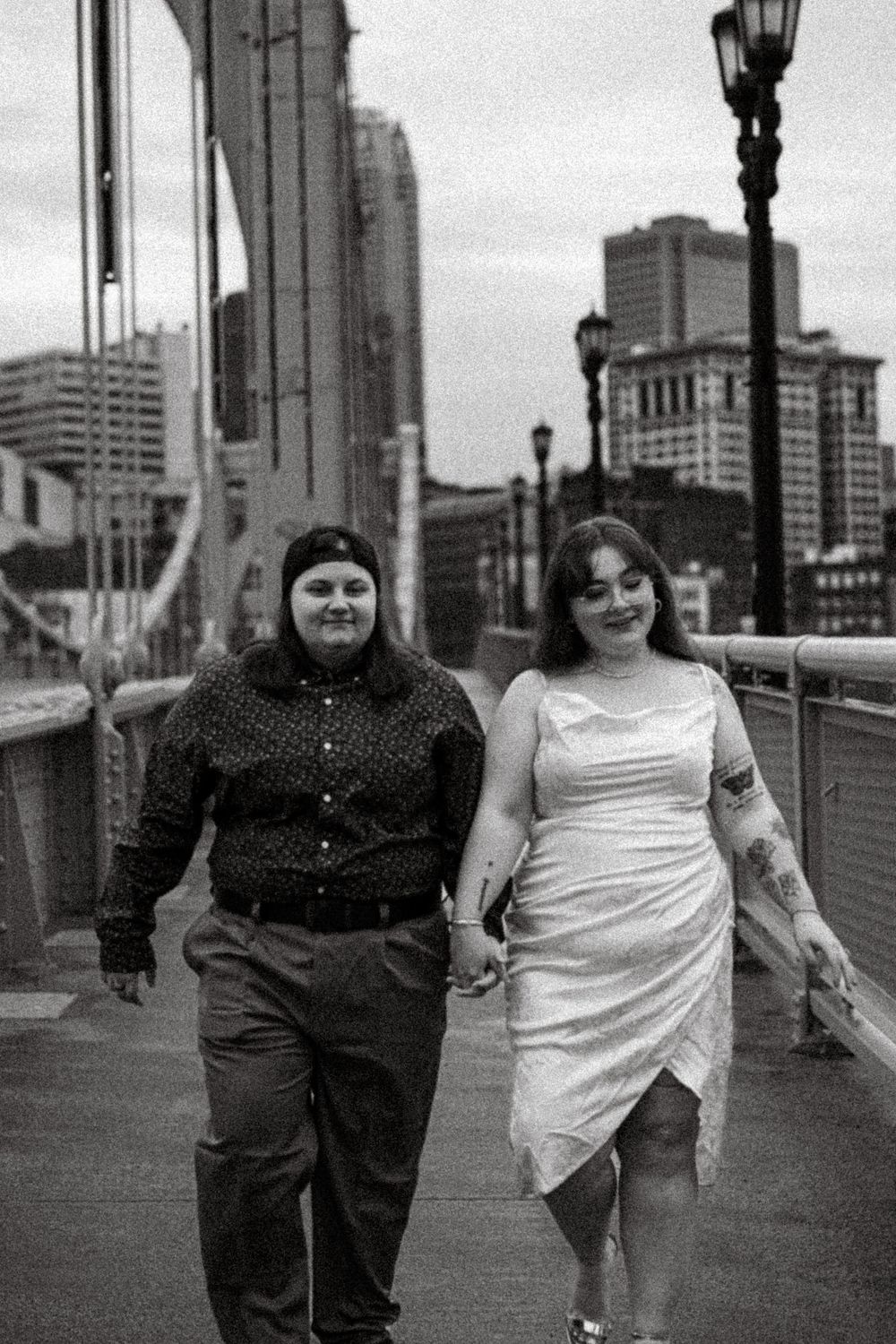 Two people walking together on a city bridge with urban buildings and lampposts visible in the background.