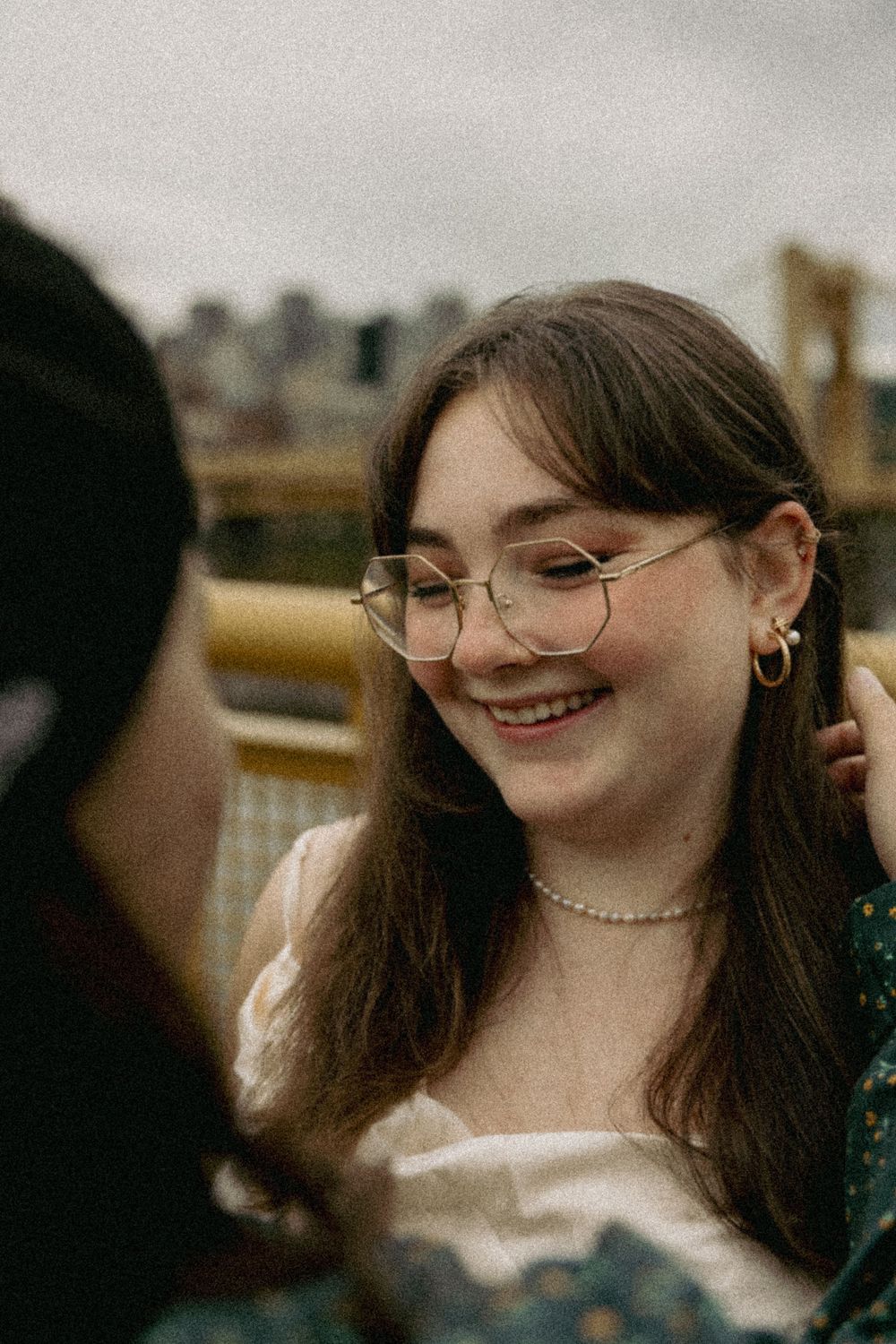 A joyful candid moment shows a person with long brown hair and round glasses smiling warmly outdoors.