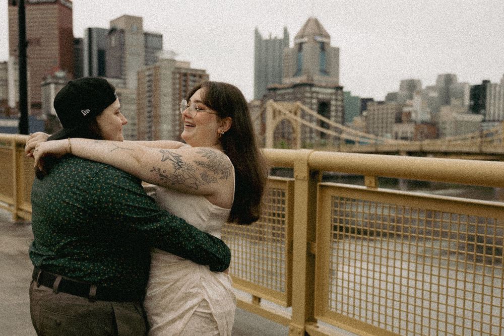A couple embraces on a rooftop deck with Pittsburgh's downtown skyline visible in the background.