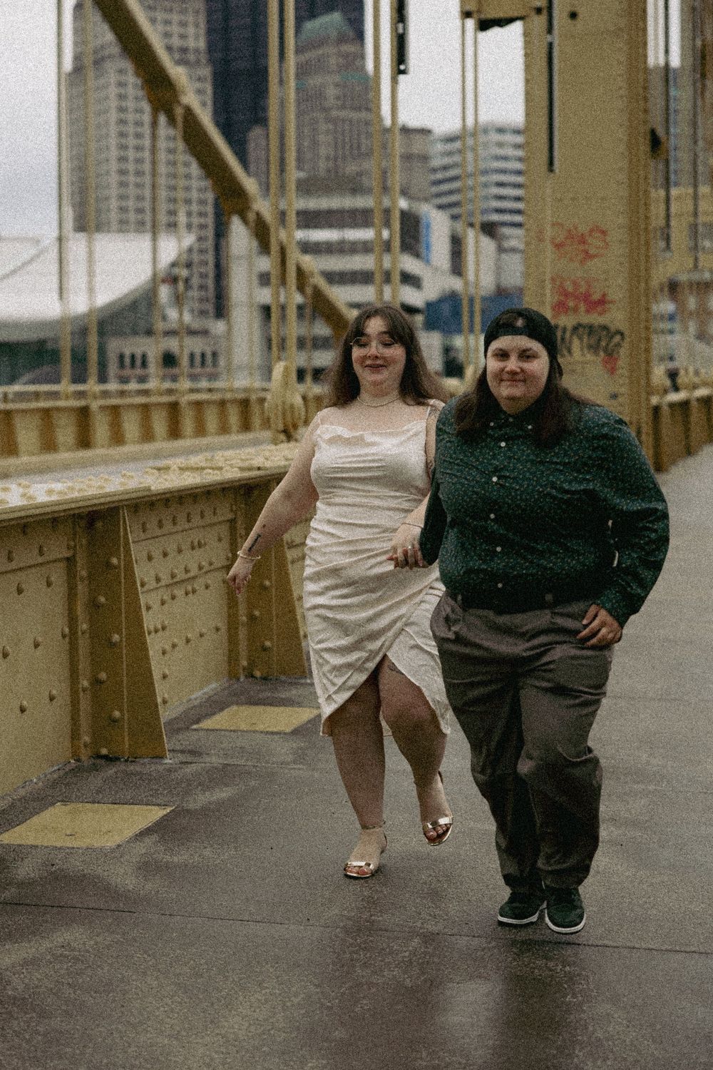 Two friends walking together on an urban bridge with city buildings visible in the background.