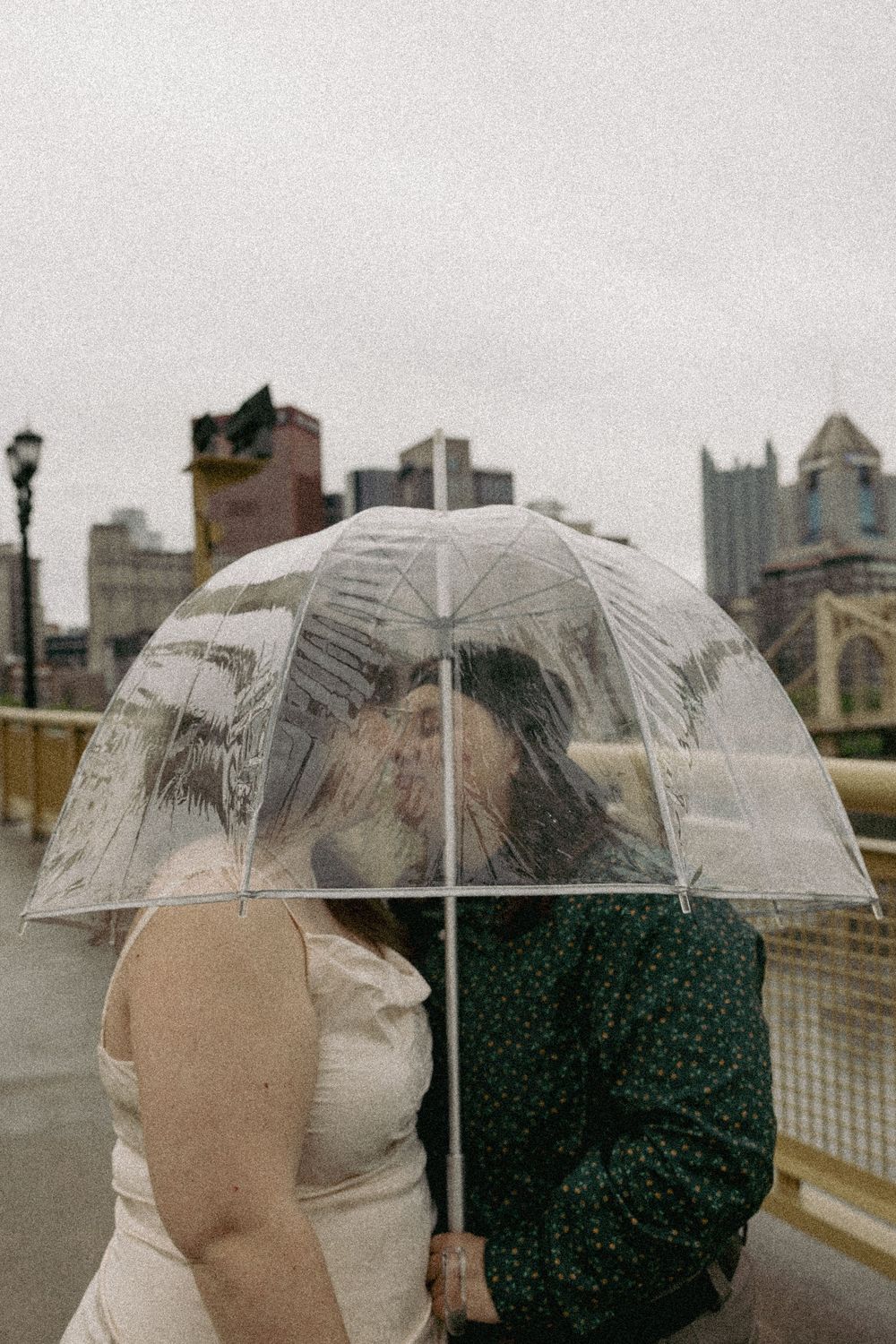 Two people share a clear umbrella on a rainy city street with buildings visible in the background.