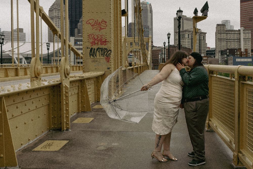 A couple in casual attire sharing a romantic moment on a yellow bridge with a city skyline behind them.