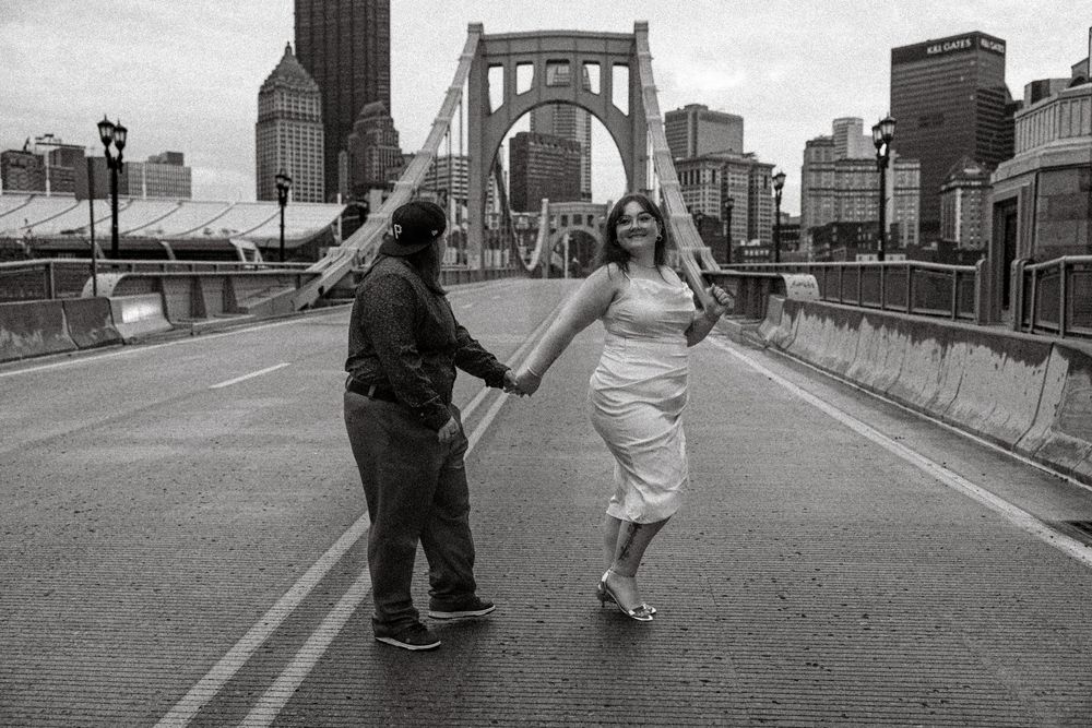 A couple holding hands while walking down a bridge with the Pittsburgh cityscape in the background.