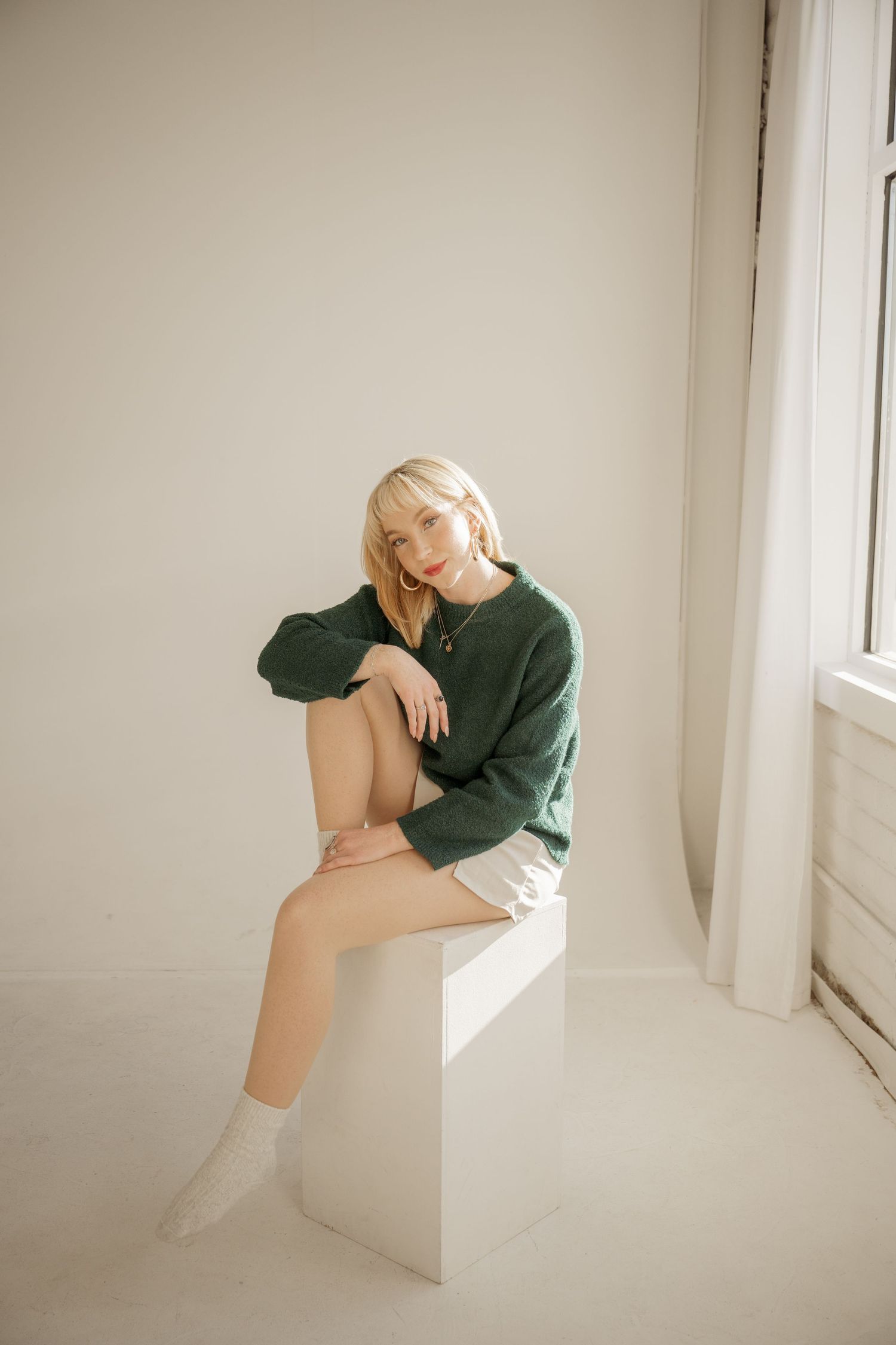 Woman in dark green sweater and white socks sits casually on white pedestal by window in minimalist setting.