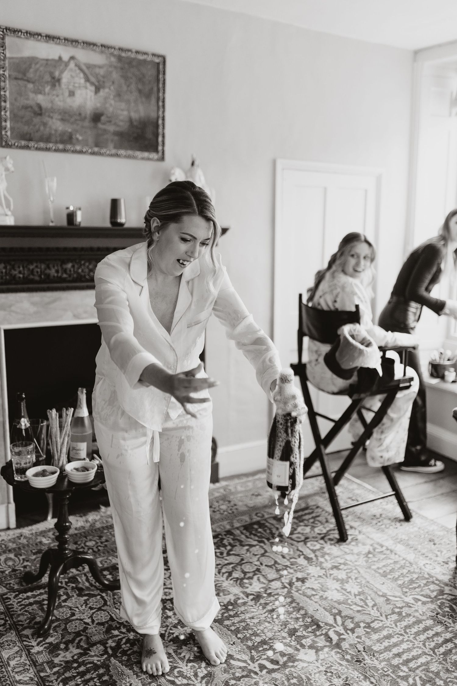 Bride in a white pjs opens champagne while getting ready in a room with a fireplace and ornate rug in Montgomery before wedding.