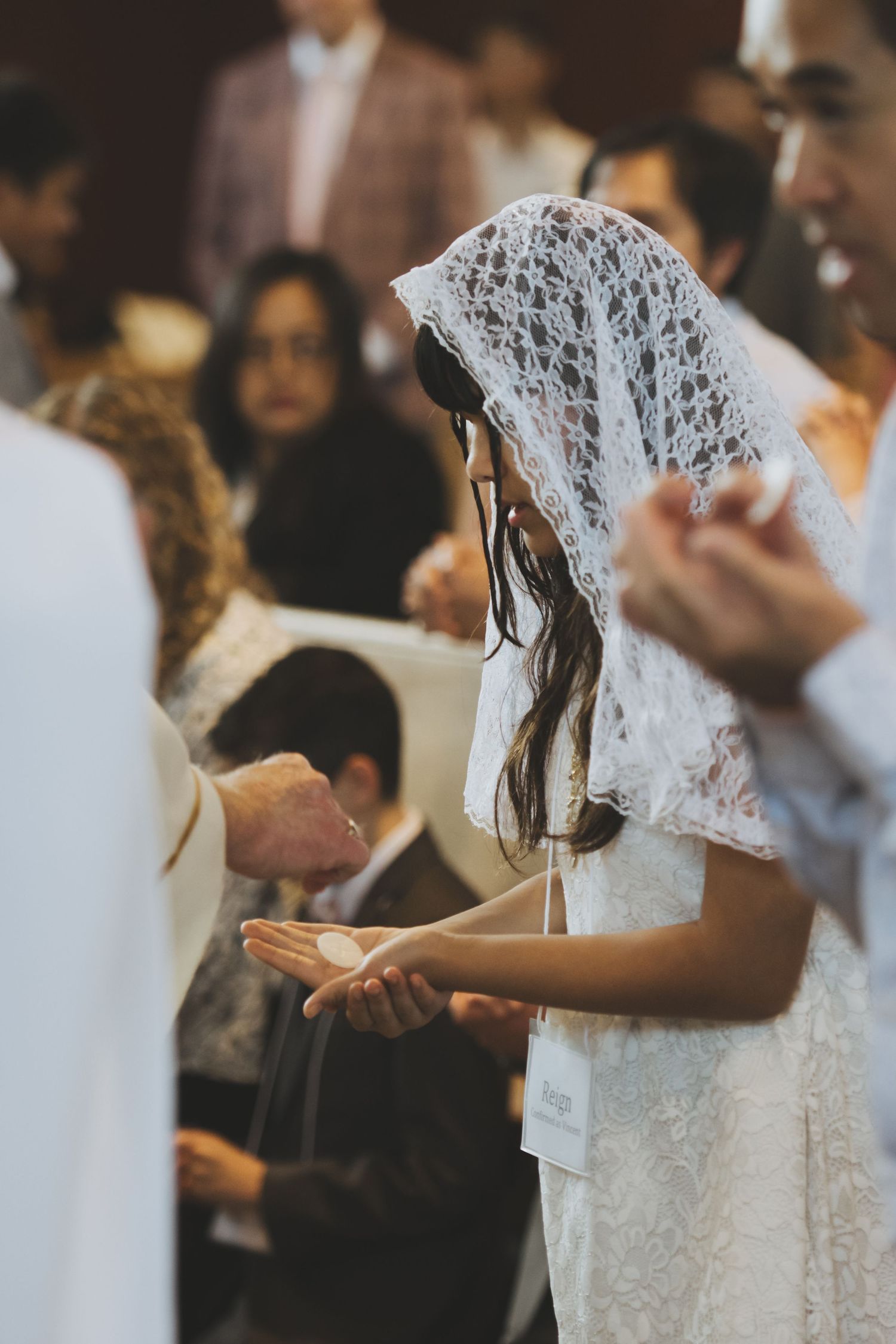 White-veiled girl wearing a lace dress holds her hands out during traditional Catholic confirmation mass.
