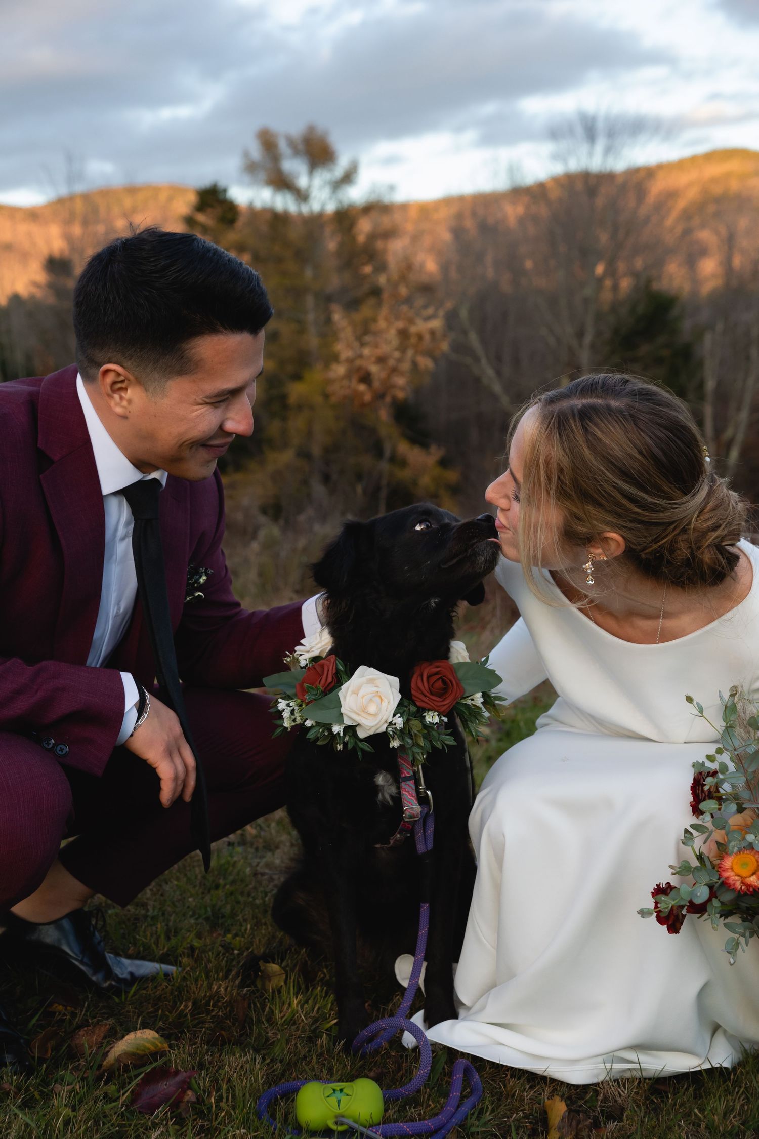 A couple in wedding attire shares a joyful moment with their black dog against an autumn mountain backdrop at sunset.