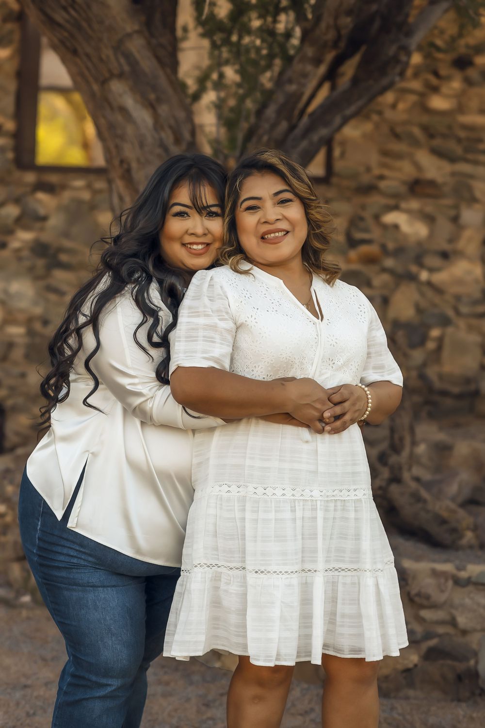 Two friends in white outfits share a joyful moment together while posing against a rustic stone wall outdoors.