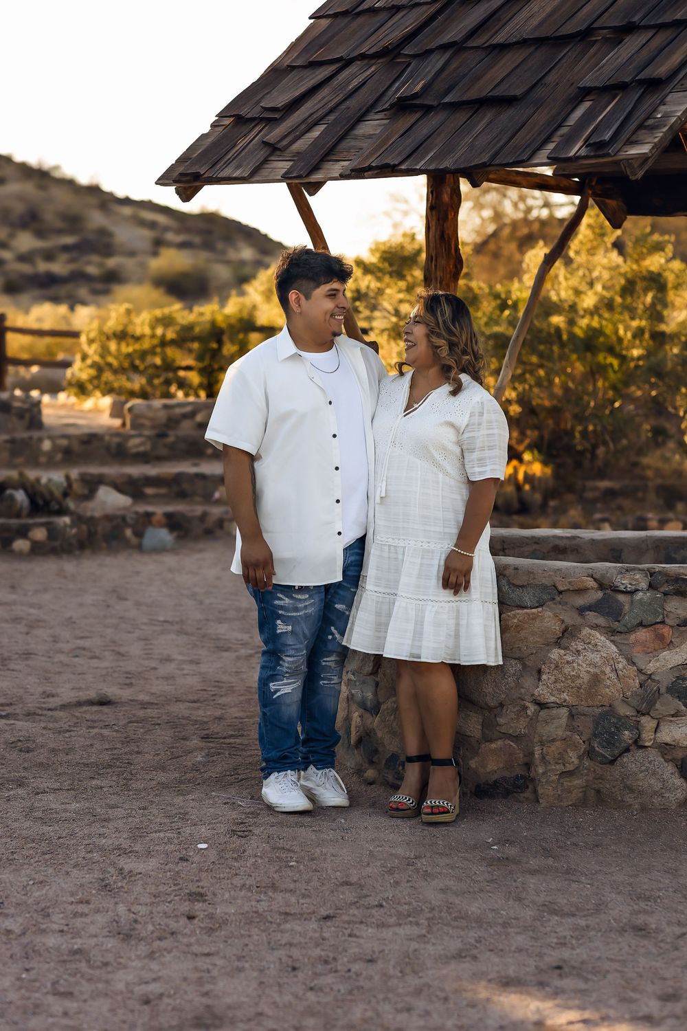 Couple in white outfits standing together under a wooden shelter during golden hour at a rustic outdoor location.